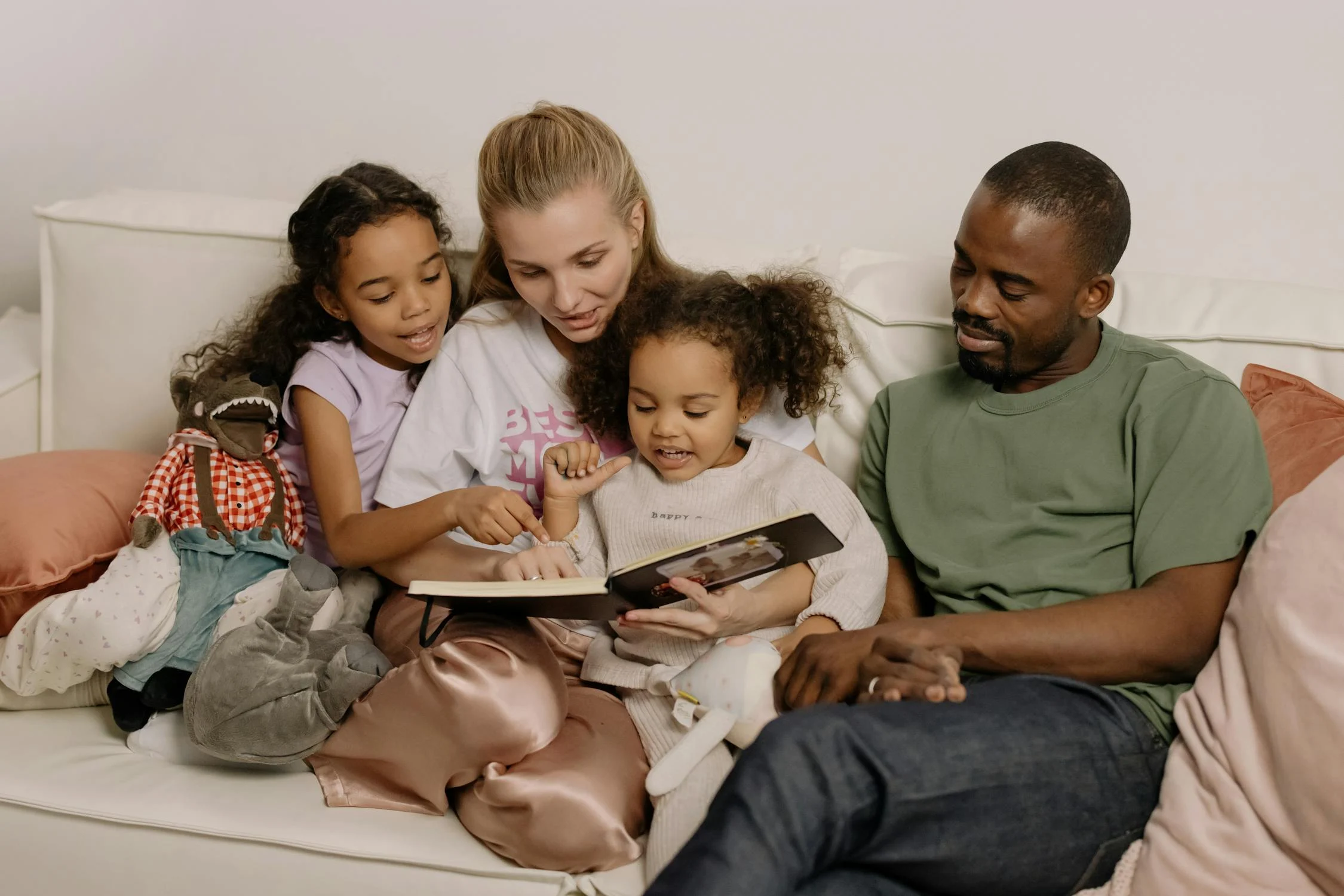 A family of four sits on a couch reading a book together; two young children are seated between an adult woman and an adult man, with a stuffed monkey toy next to them.